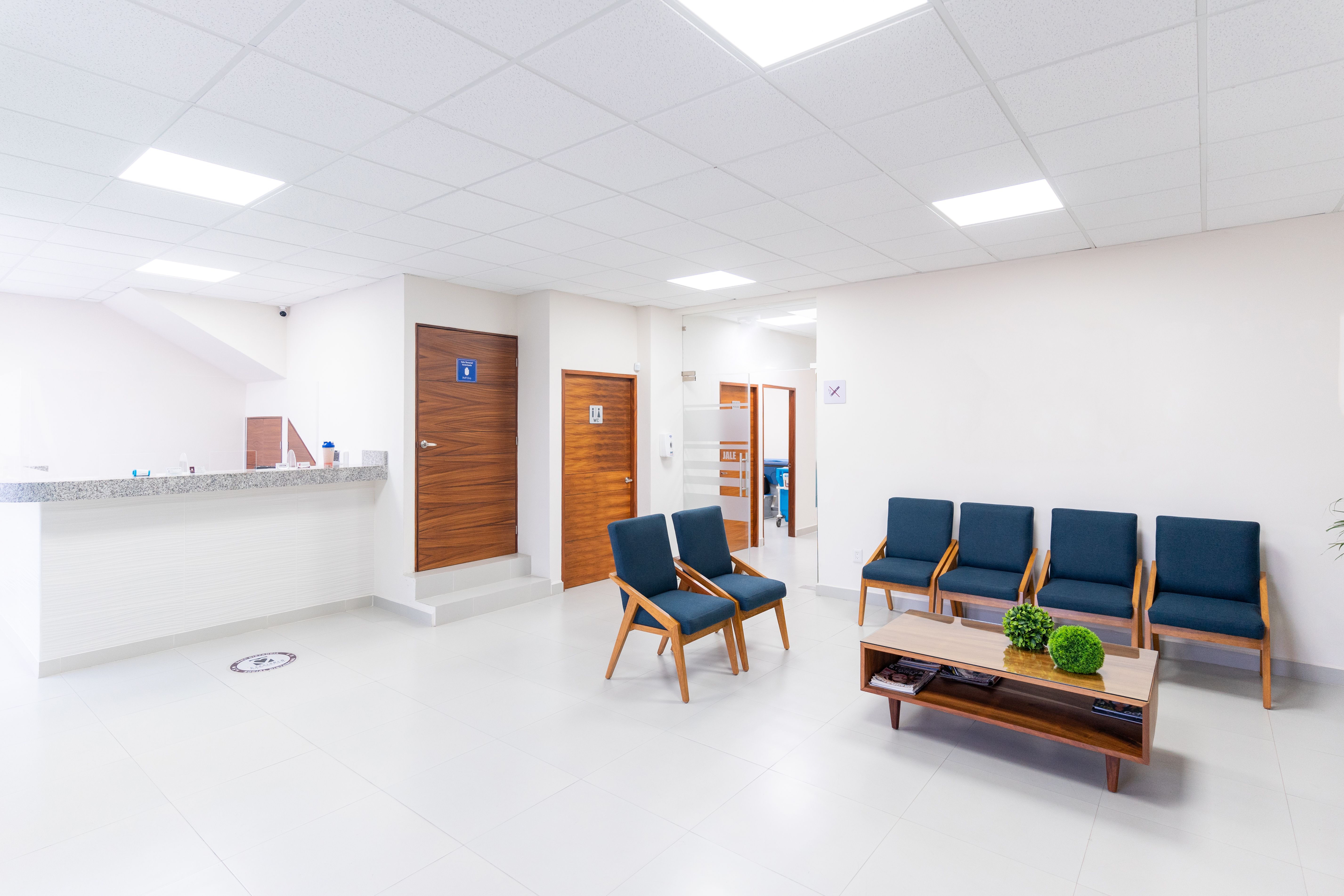 Empty clinic waiting area with a reception counter, navy upholstered chairs, and a low wooden table with magazines under bright ceiling lights